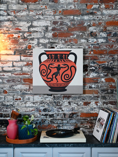 This art print depicts a clay vase with a Herculean figure fighting off giant snakes and is accented by historically-fitting patterns like narrative figures and meanders. The print is displayed against a brick wall in a modern home.