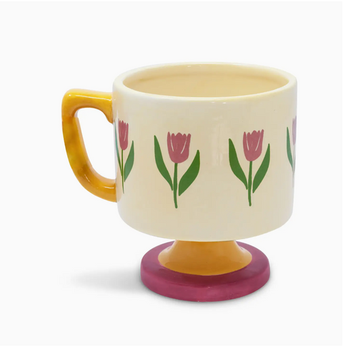 A ceramic mug with an off-white body, yellow handle, and pink tulip patterns on the side, sitting on a pink pedestal base.