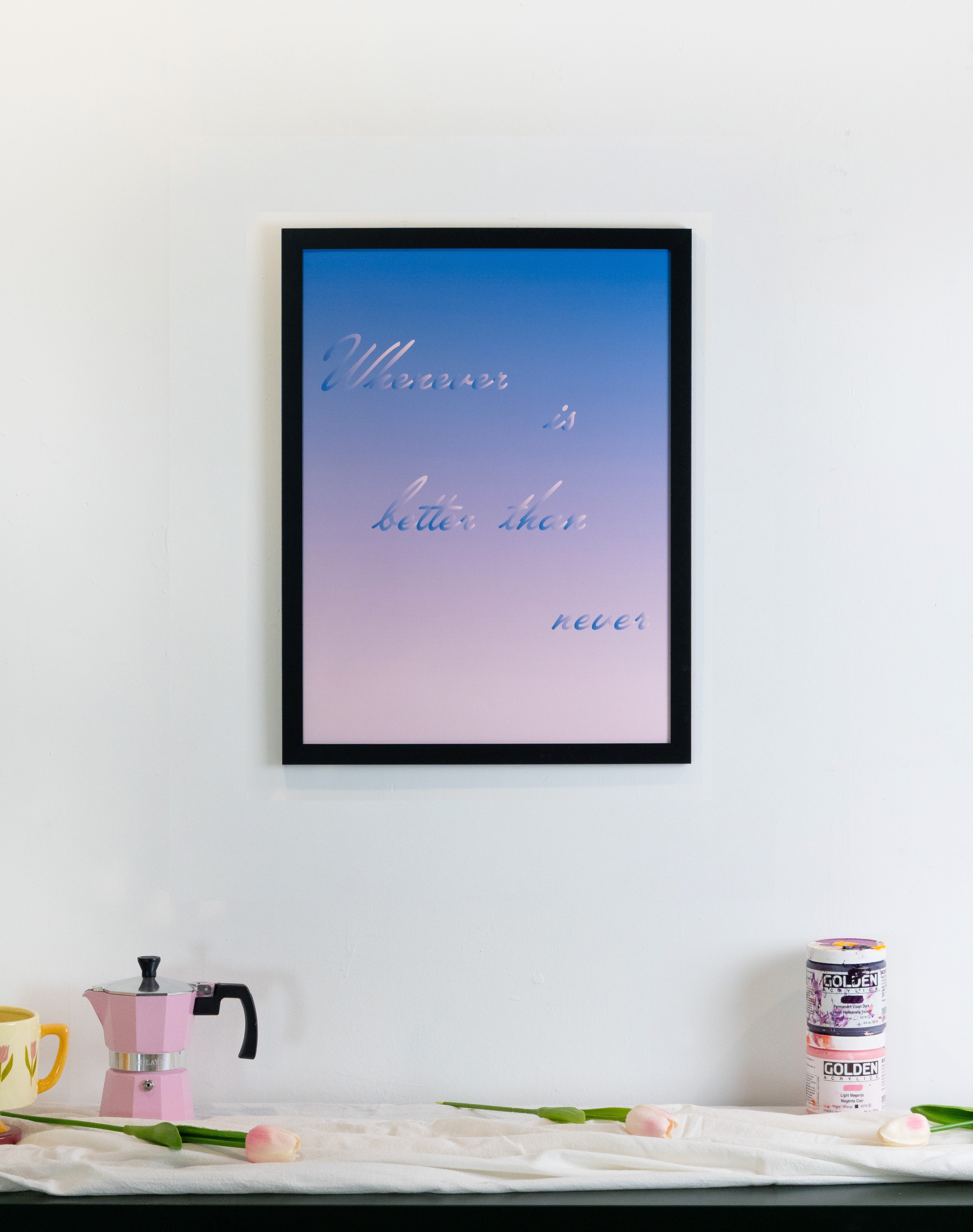 Framed artwork with a gradient background and text on a wall above a table with a coffee maker and flowers.
