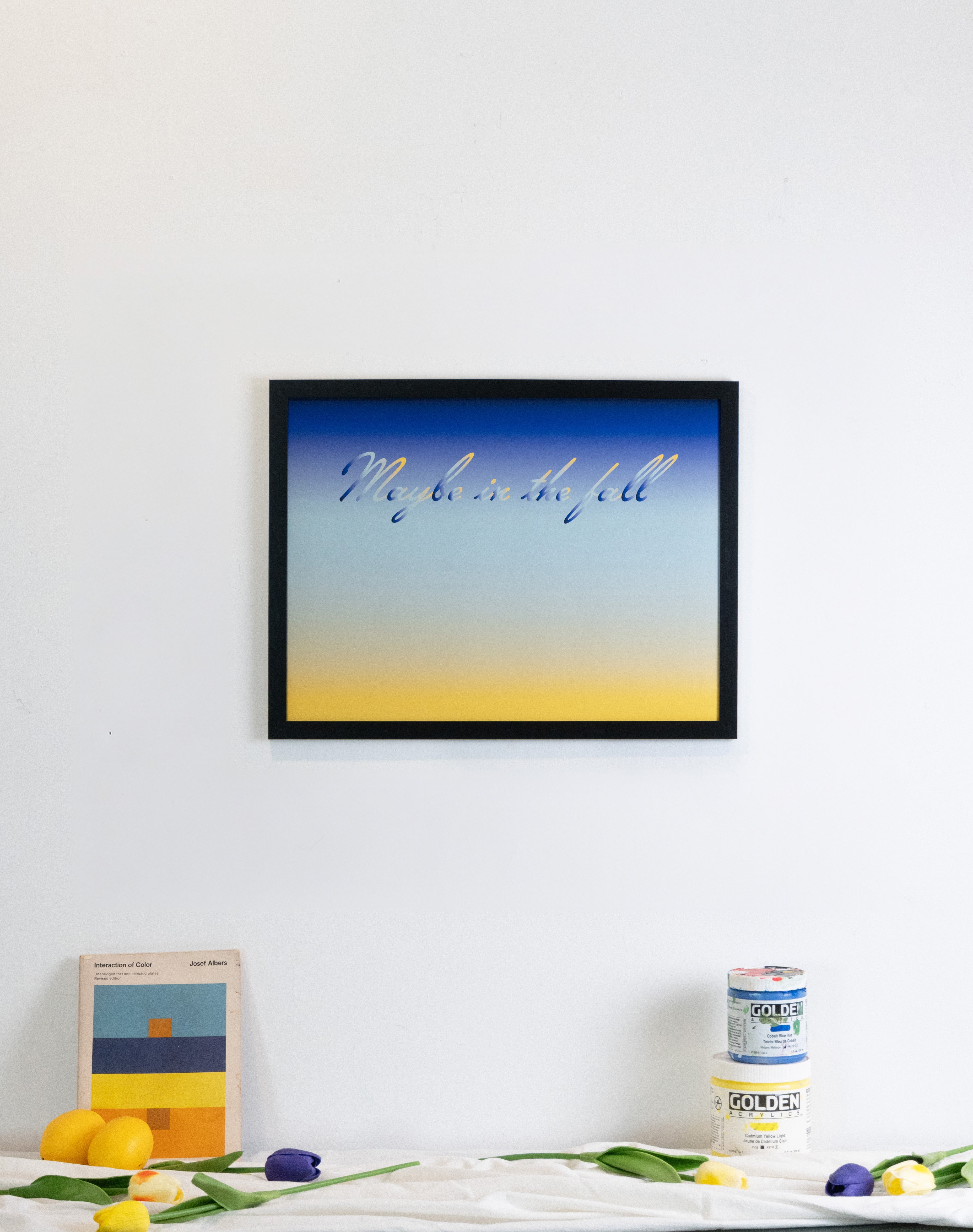 Framed artwork with 'Maybe in the fall' text on a gradient background, displayed on a wall with decor items.