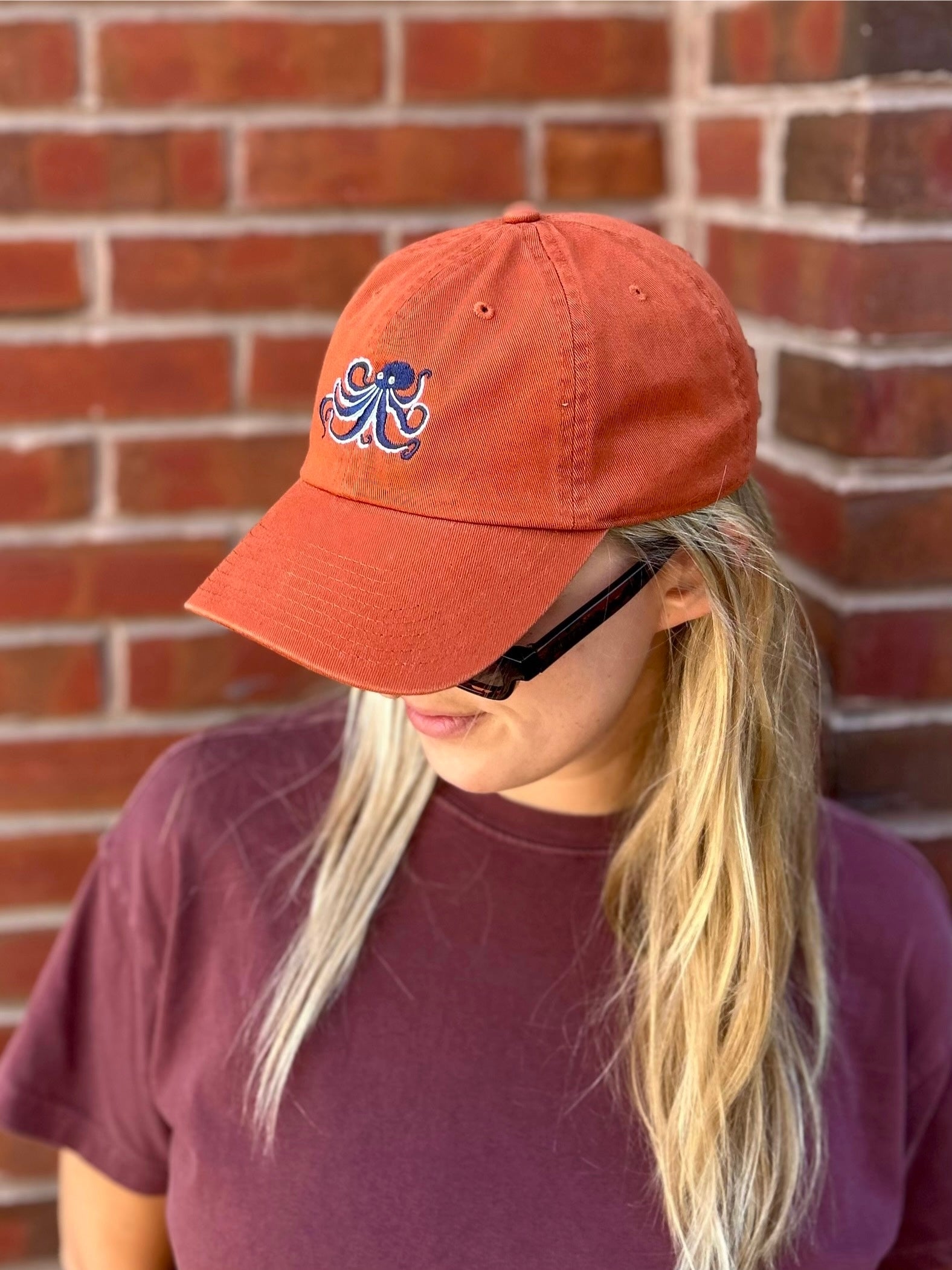 Person wearing an orange cap with an octopus embroidery design, standing against a brick wall.