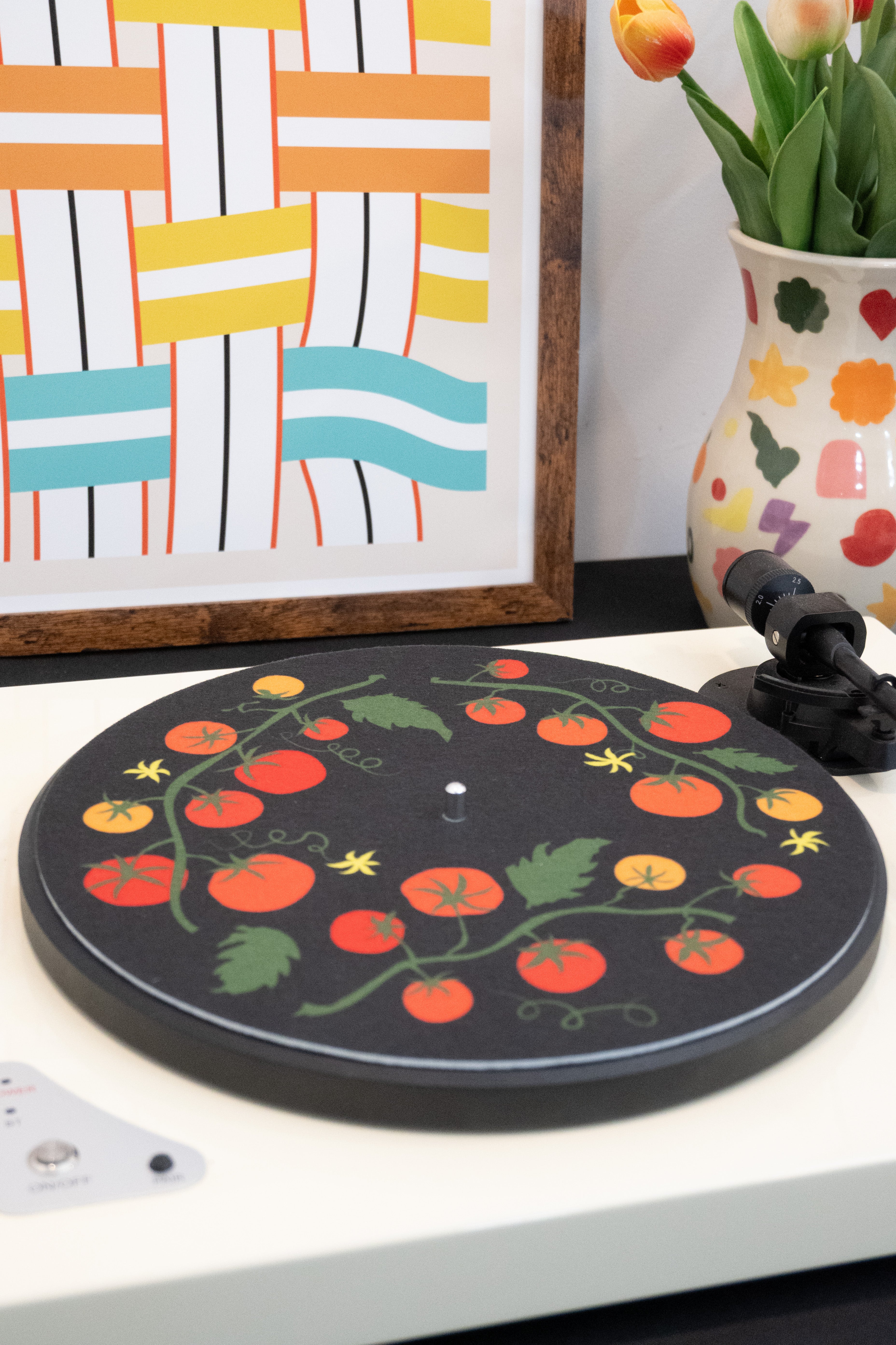 Decorative turntable with tomato design on a white surface, framed artwork in the background.