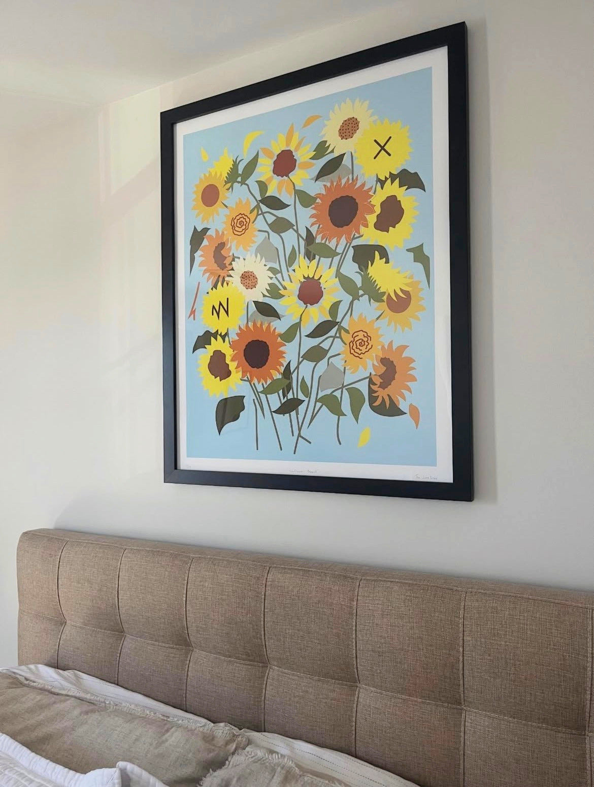 Framed artwork of contemporary sunflowers with a powder blue background on a wall above a bed.