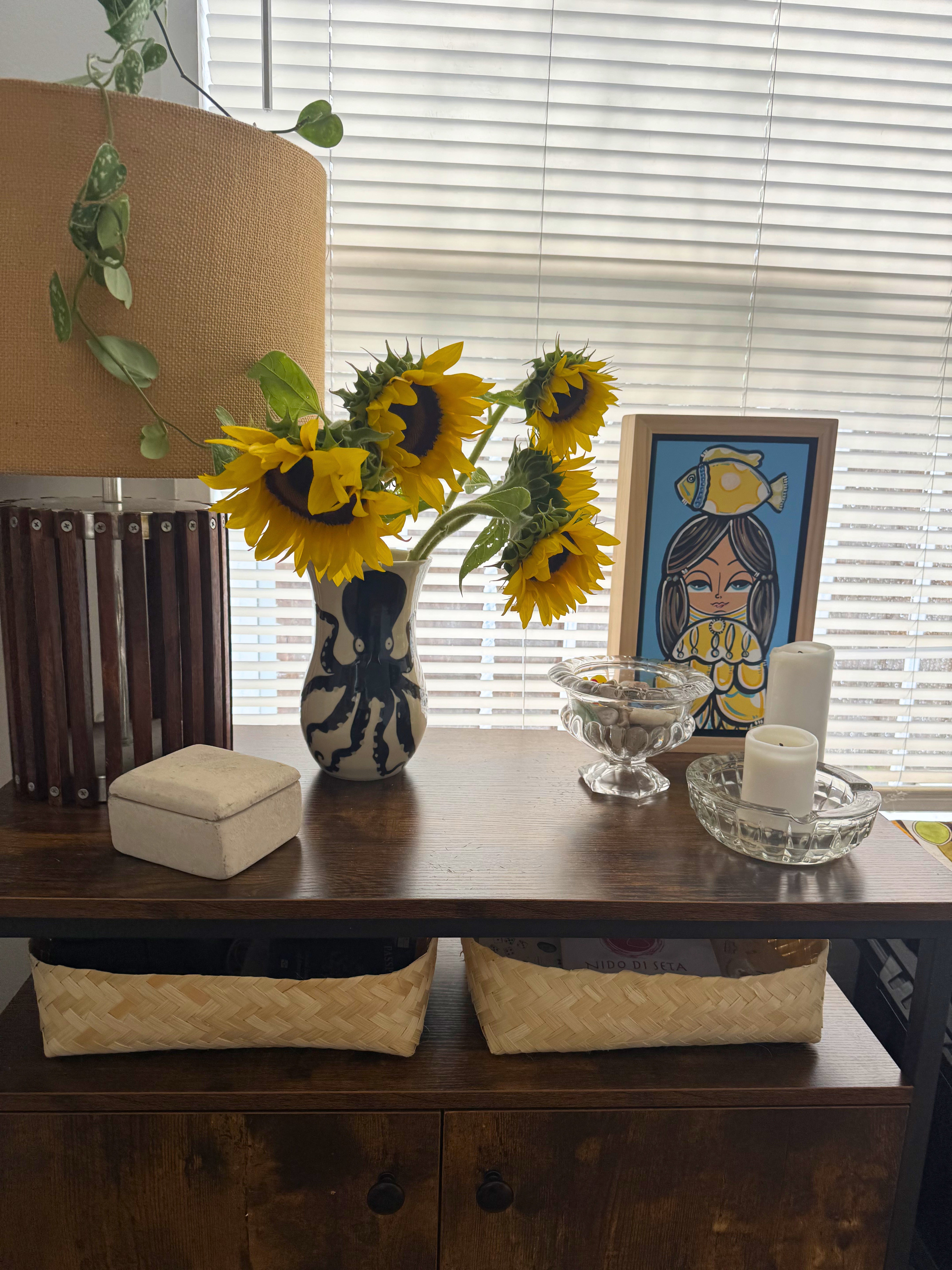 Decorative table with sunflowers, a handpainted octopus vase, and a painting in a room with drawn blinds.