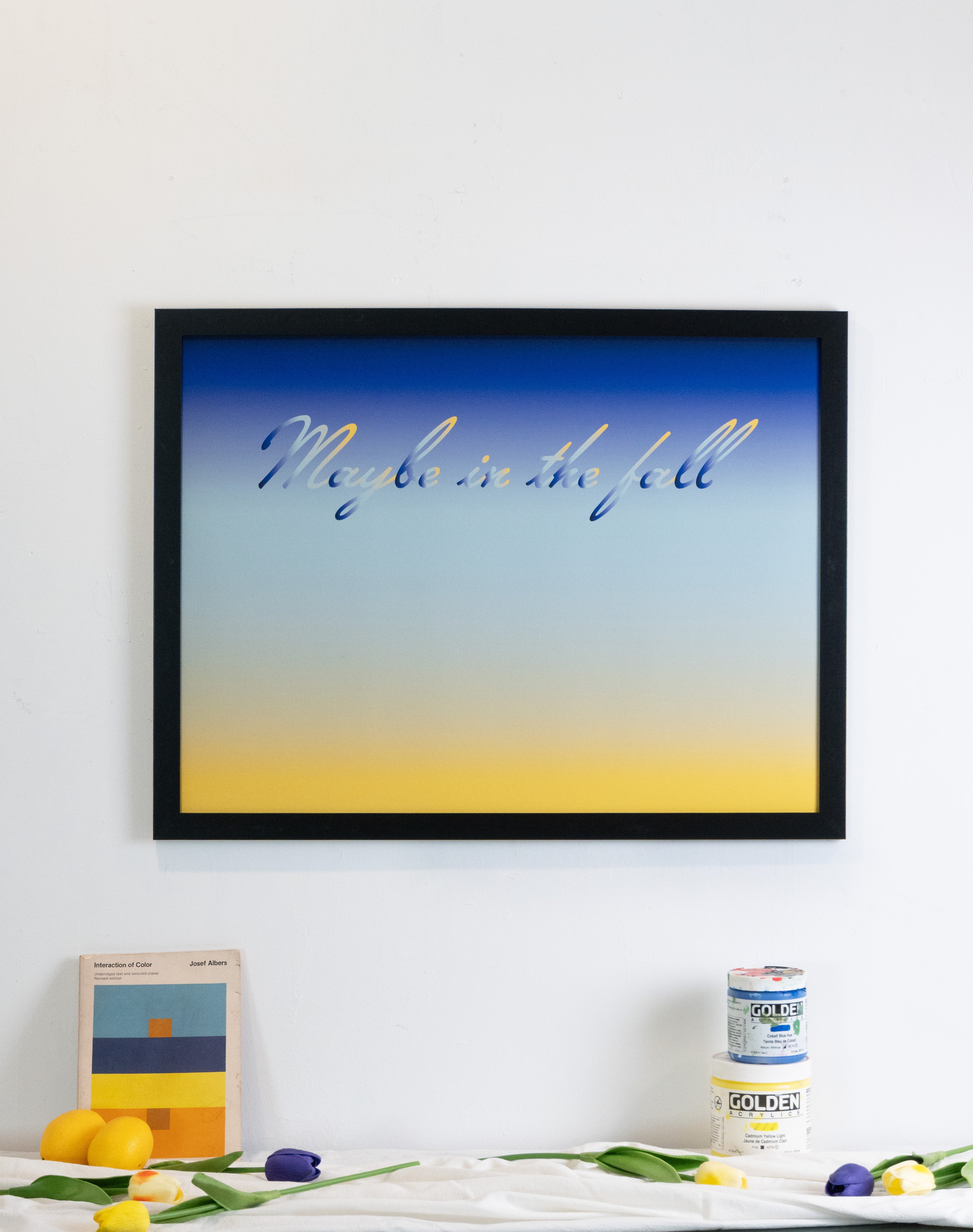 Framed artwork with 'Maybe is the fall' text on a blue and yellow gradient background, displayed on a wall and styled with decorative items