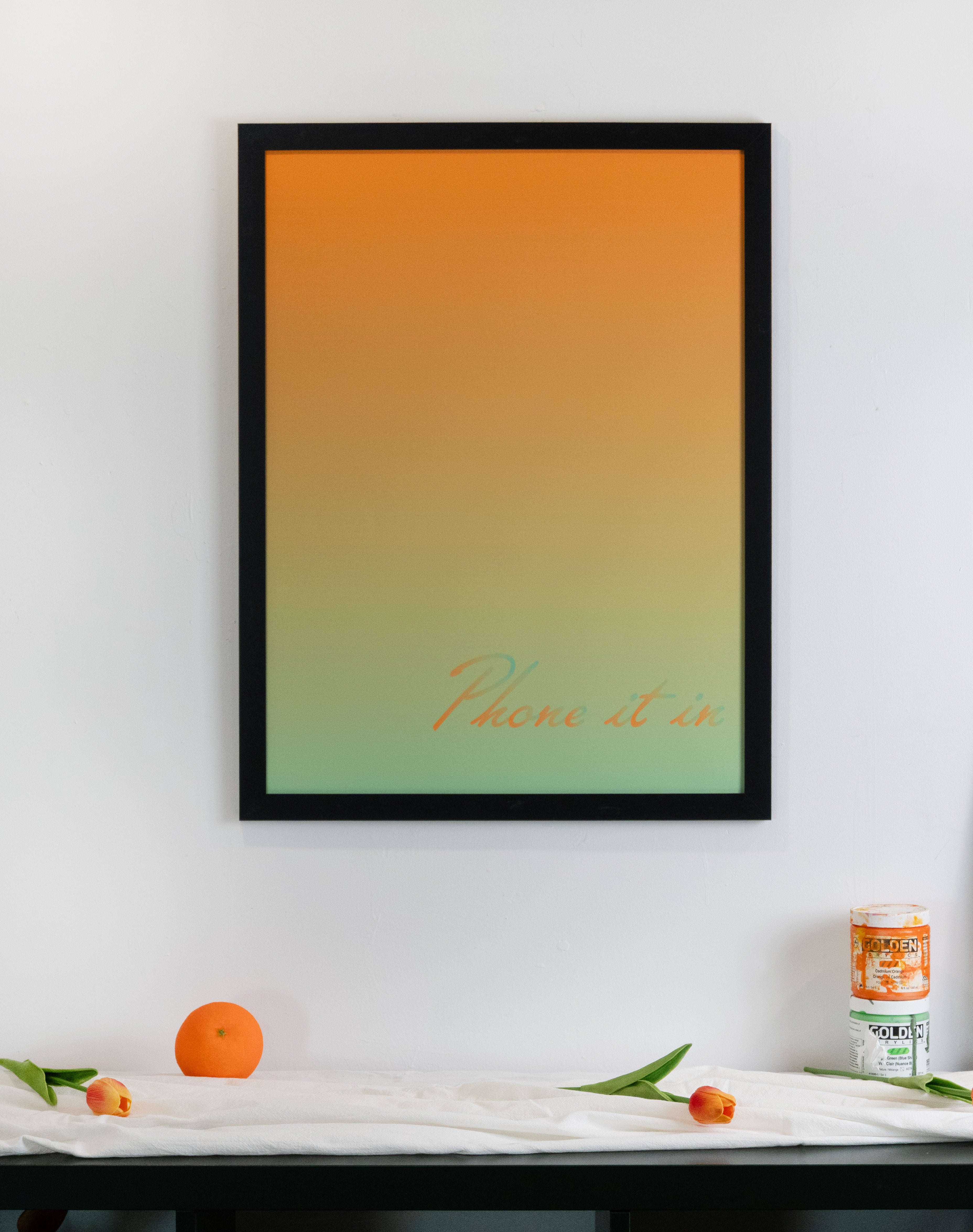 Framed artwork with a green and orange gradient design and text on a wall above a table with decorative items