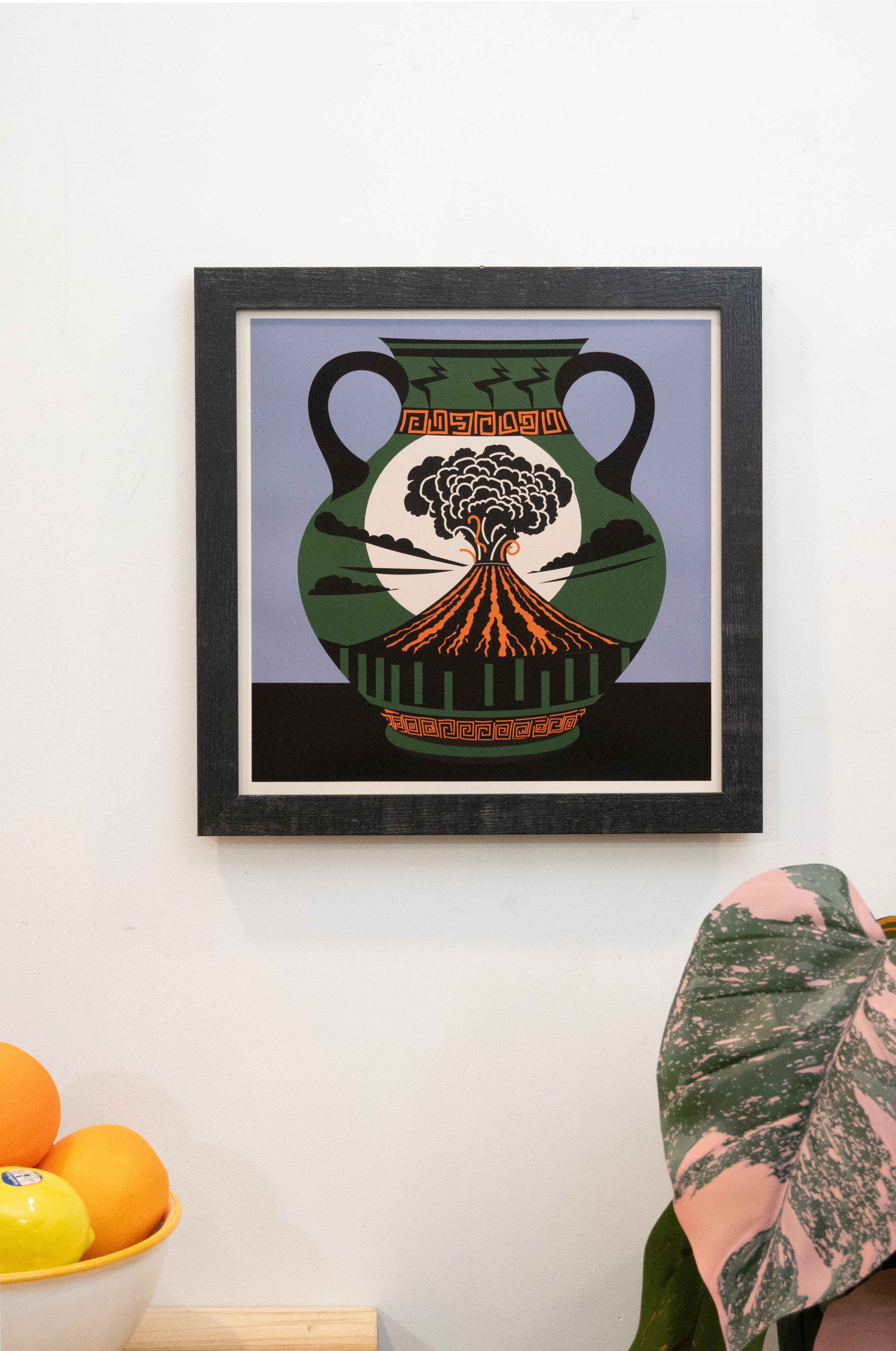 Framed art print with with volcano vase design with black and periwinkle background with white border displayed on a wall.
