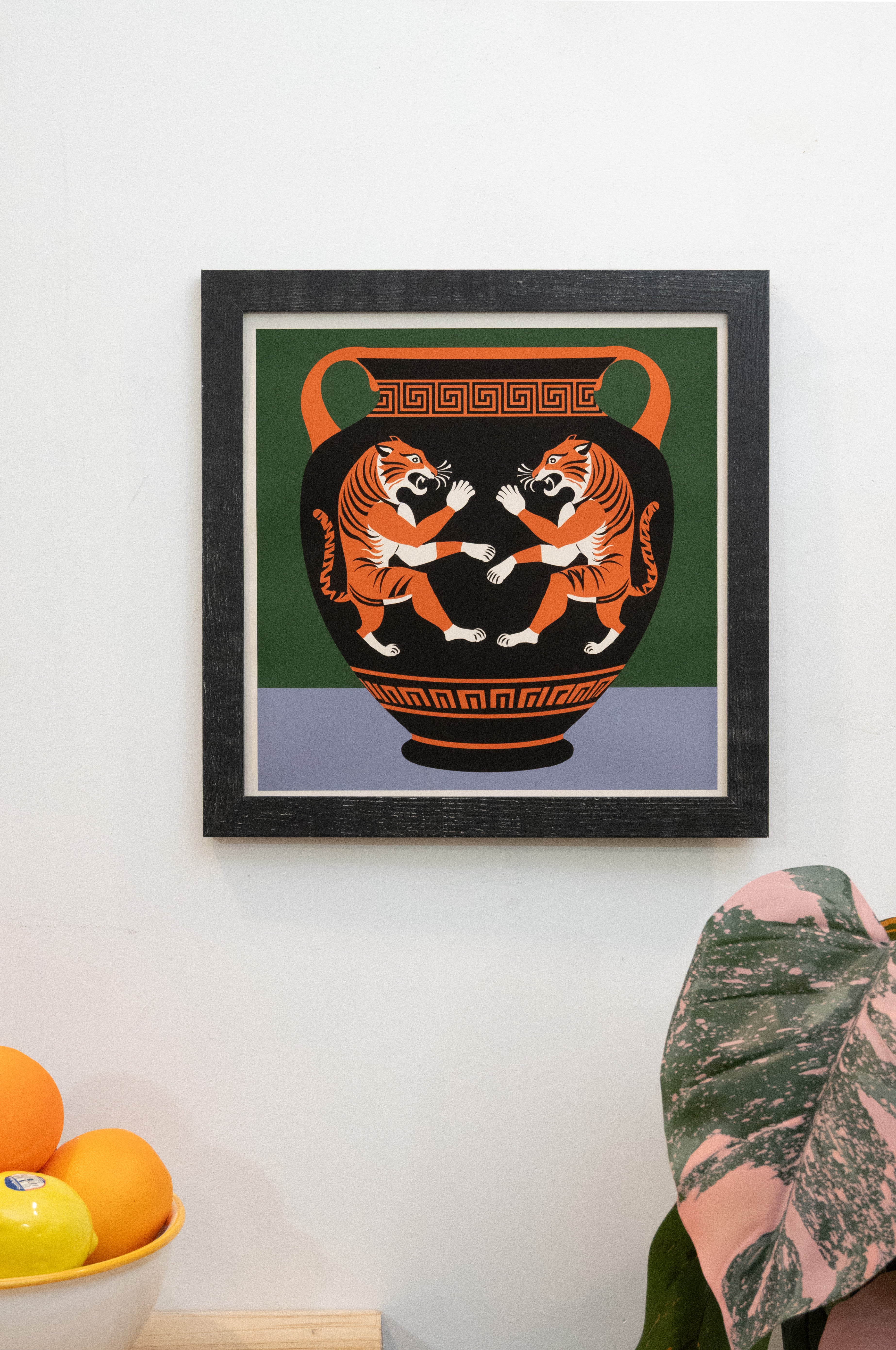 Framed artwork of two tigers on a vase against a green and periwinkle background and white border displayed on a wall.