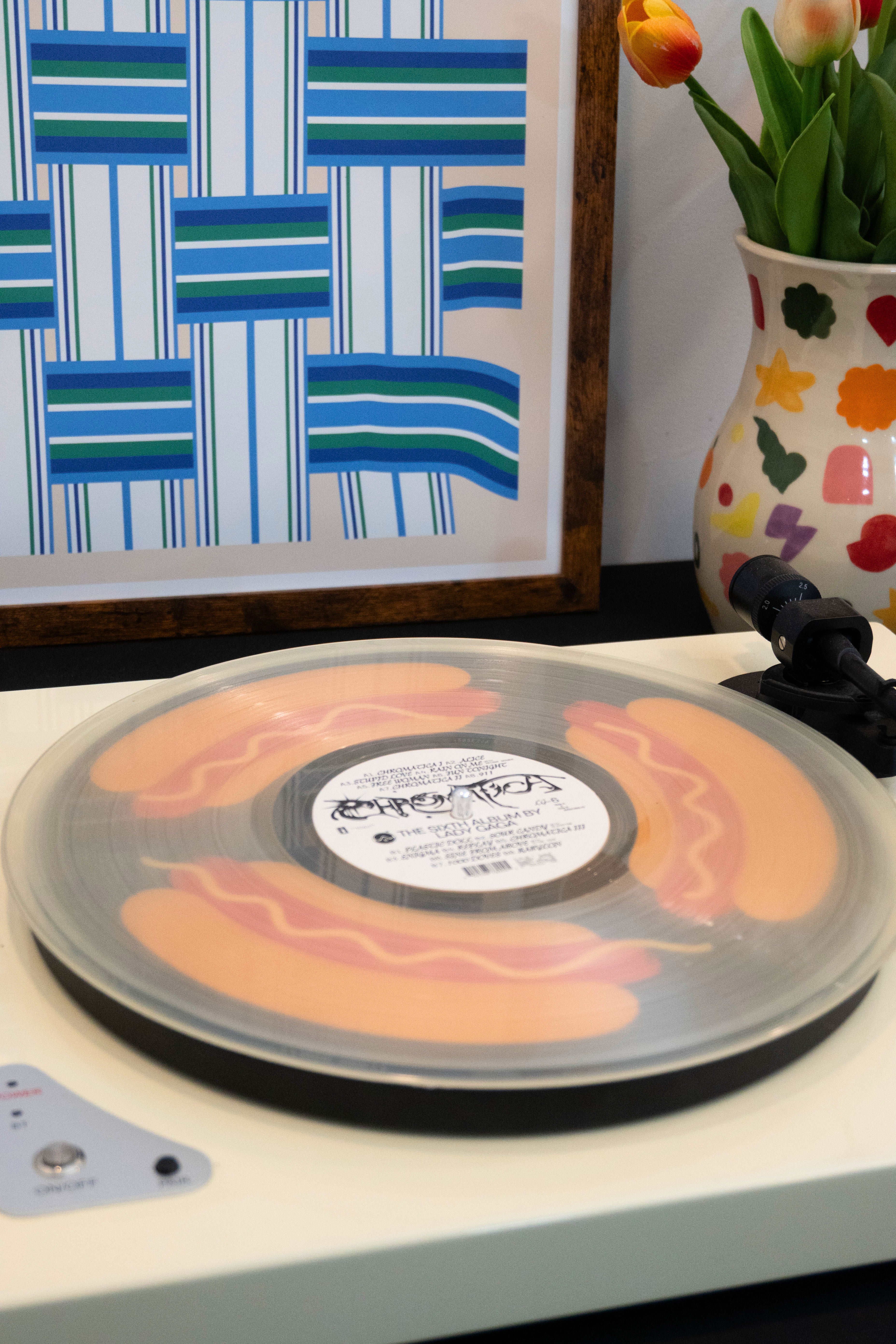 Record player with a transparent vinyl record on a turntable with a hot dog slipmat below it, next to a framed artwork and vase with flowers.