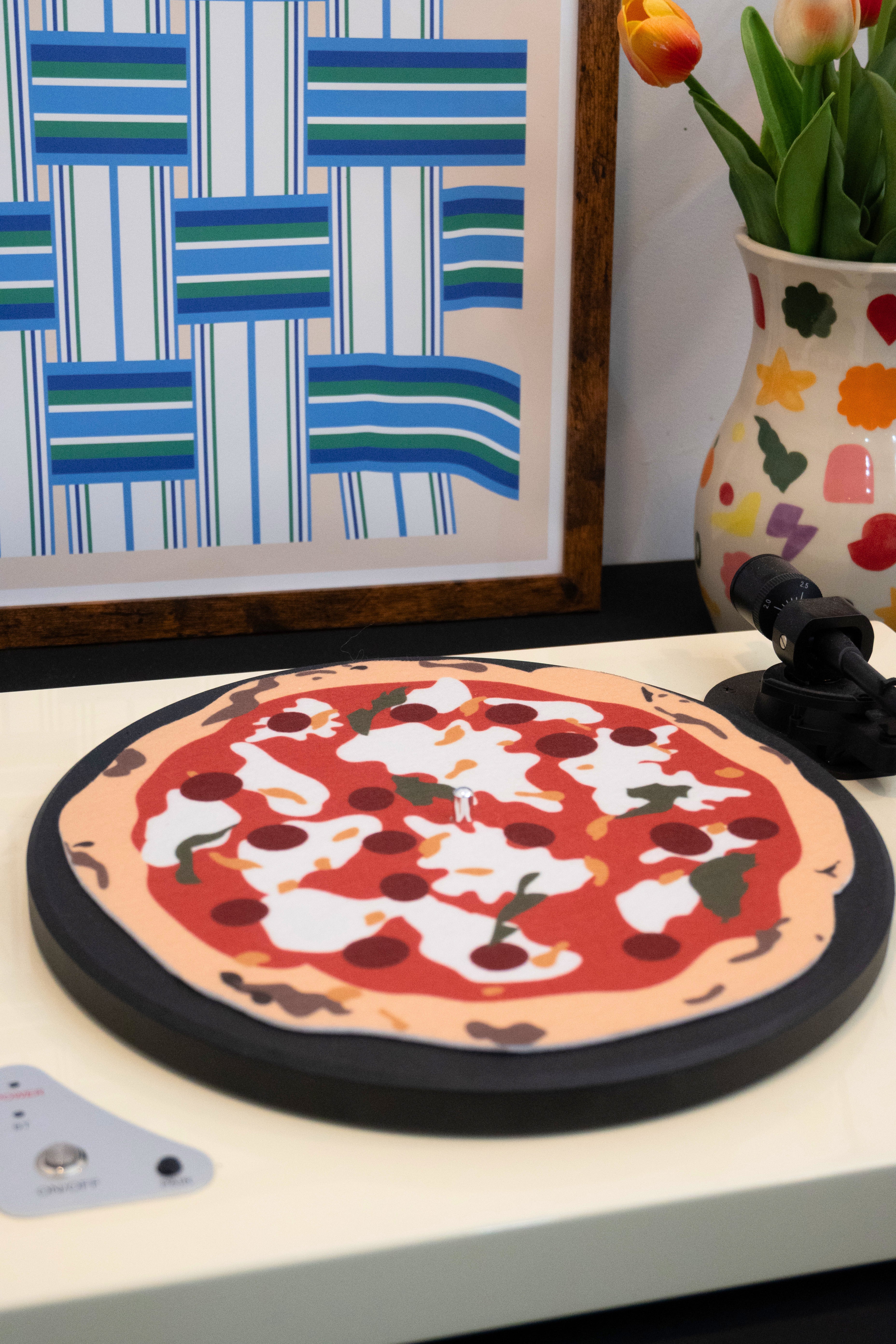 Decorative pizza slipmat on a turntable with a colorful vase and framed artwork in the background.