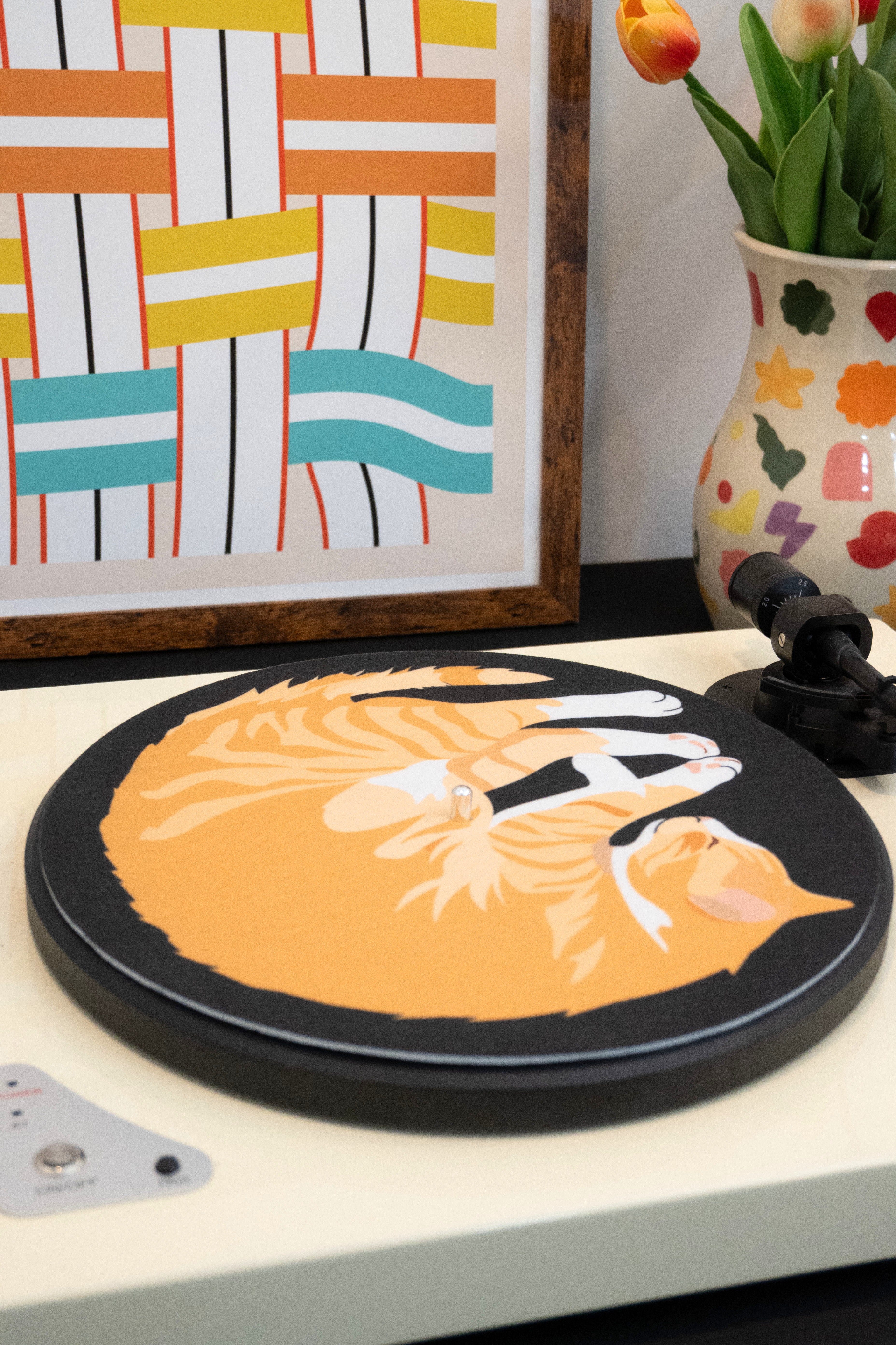 Decorative record player slipmat with a cat design on the turntable styled with colorful abstract painting and vase with tulips.