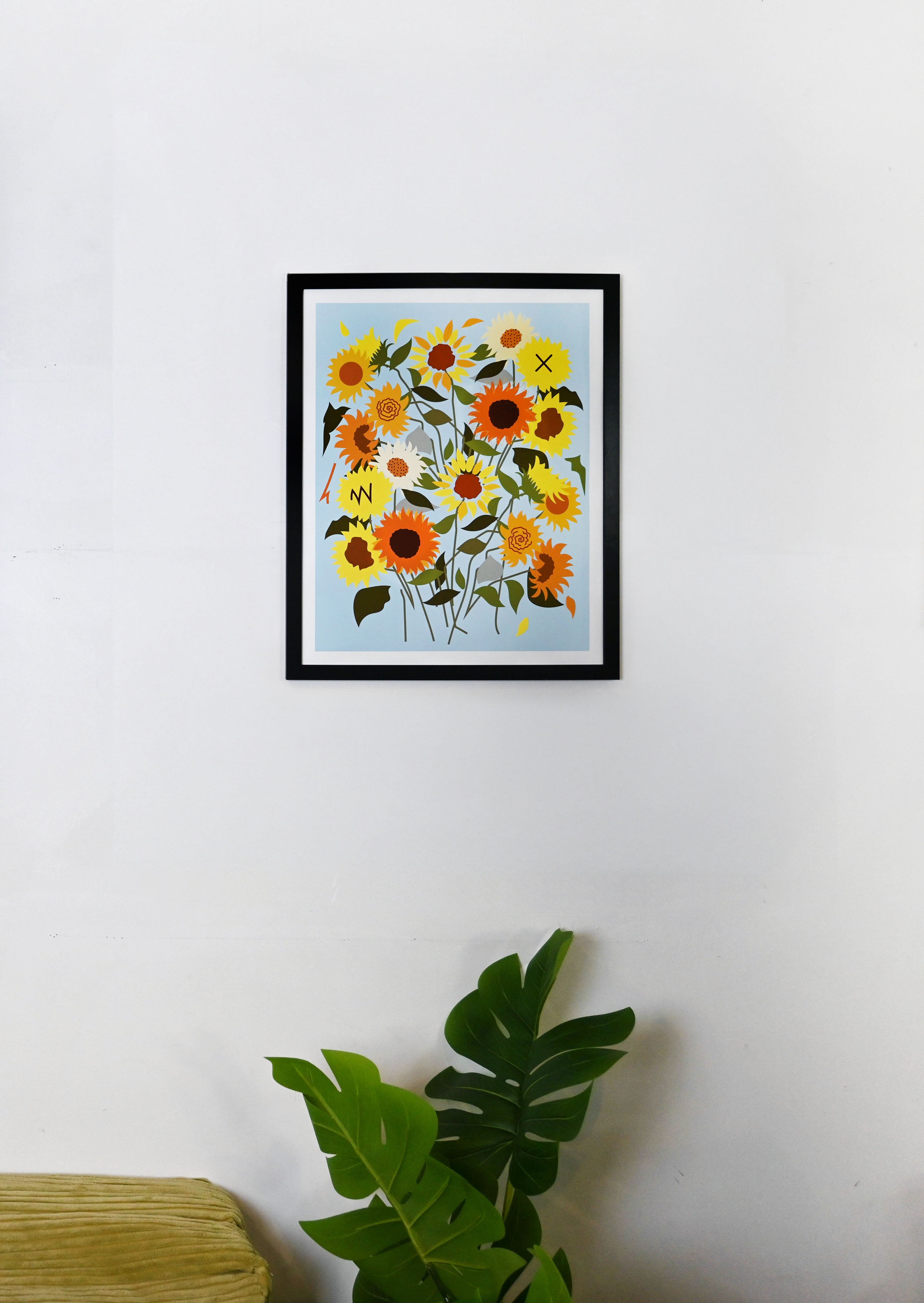 Framed artwork of sunflowers on a white wall with a plant in the foreground.