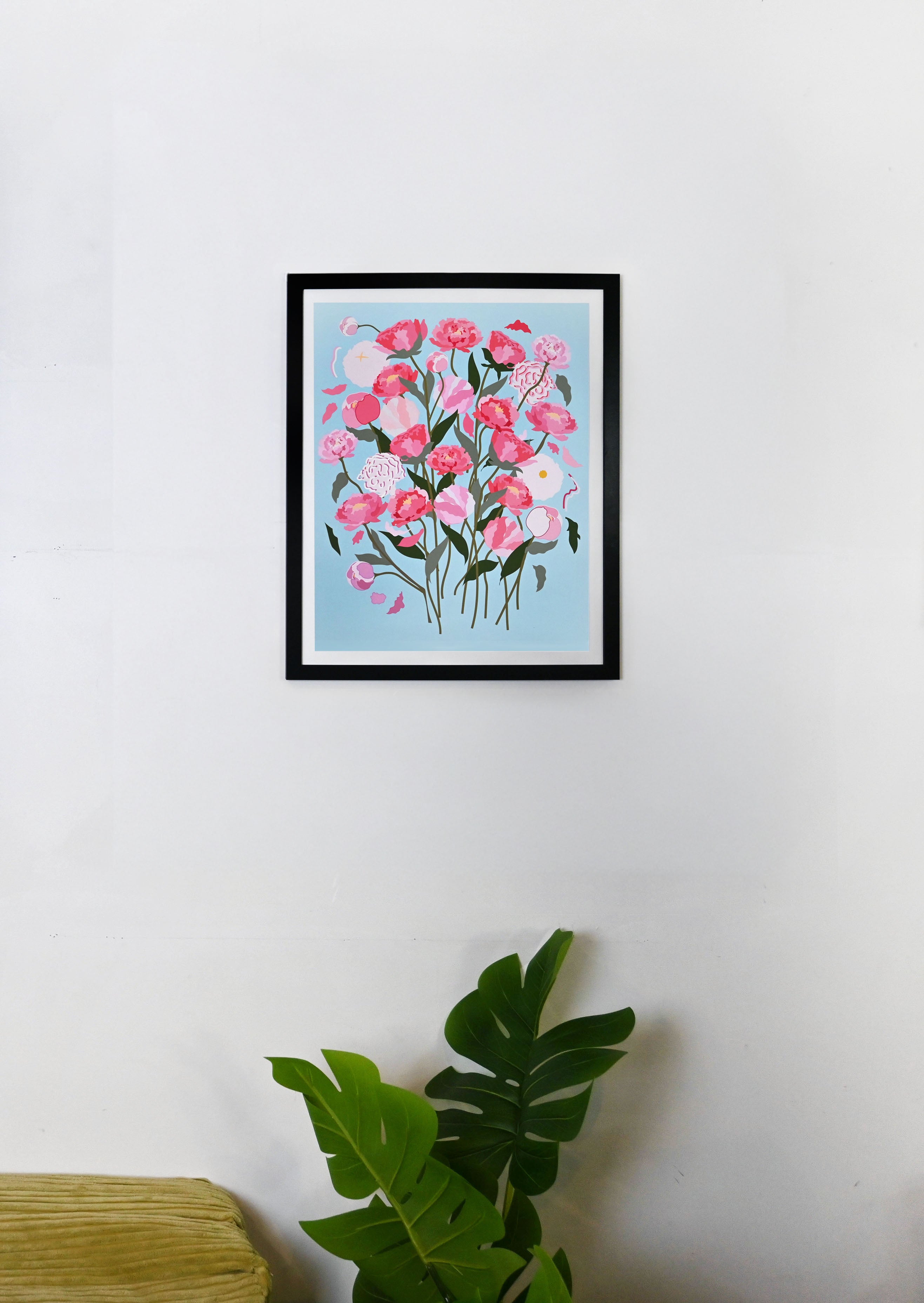 Framed artwork of pink peony flowers on a light blue background, displayed on a white wall.