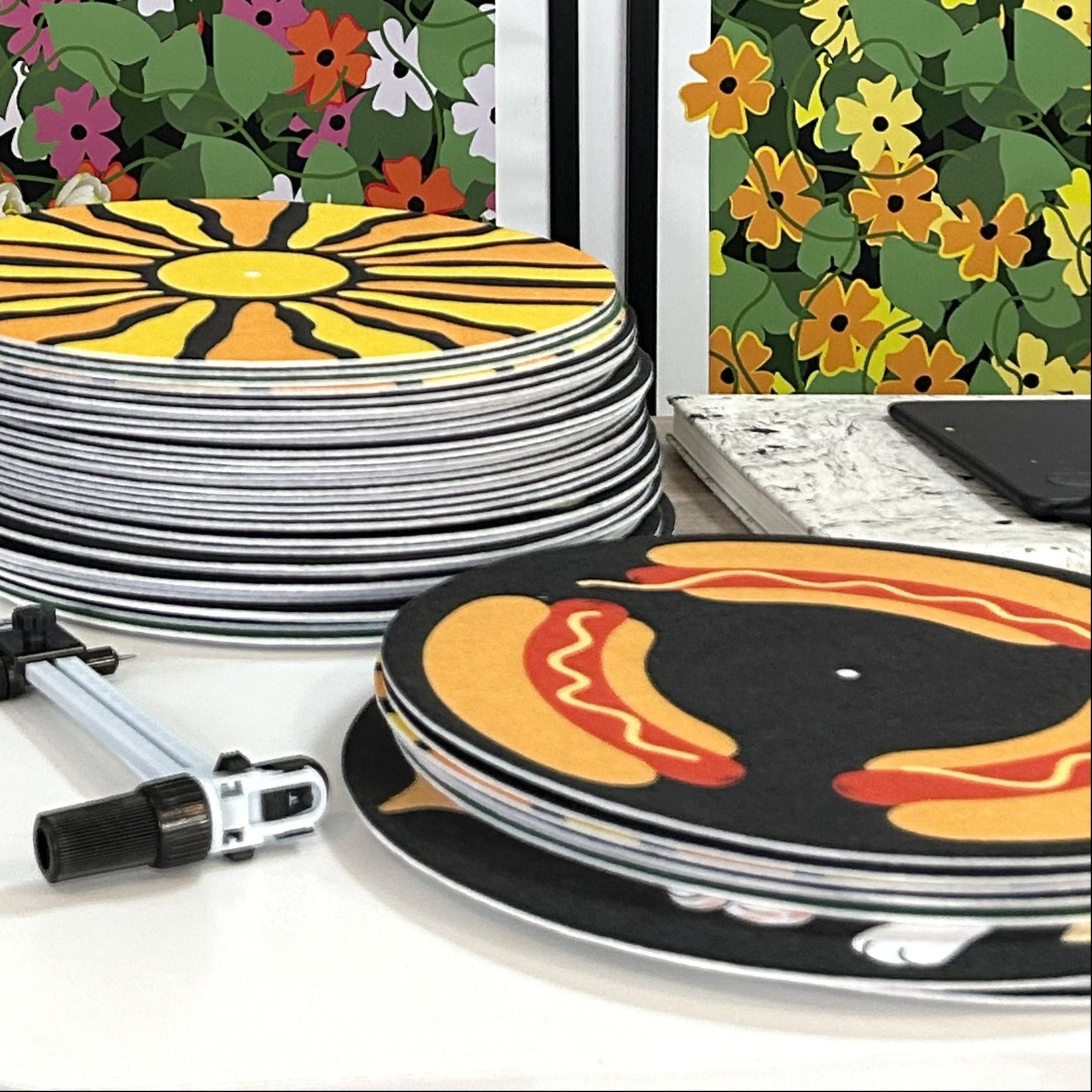 Stack of colorful turntable slipmats including sun and hot dog designs and placed in front of colorful floral wall art