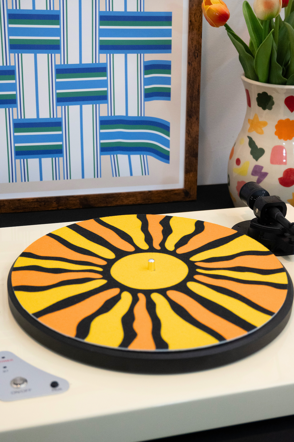 Colorful sunburst design record player slipmat on a turntable with framed art and vase in the background