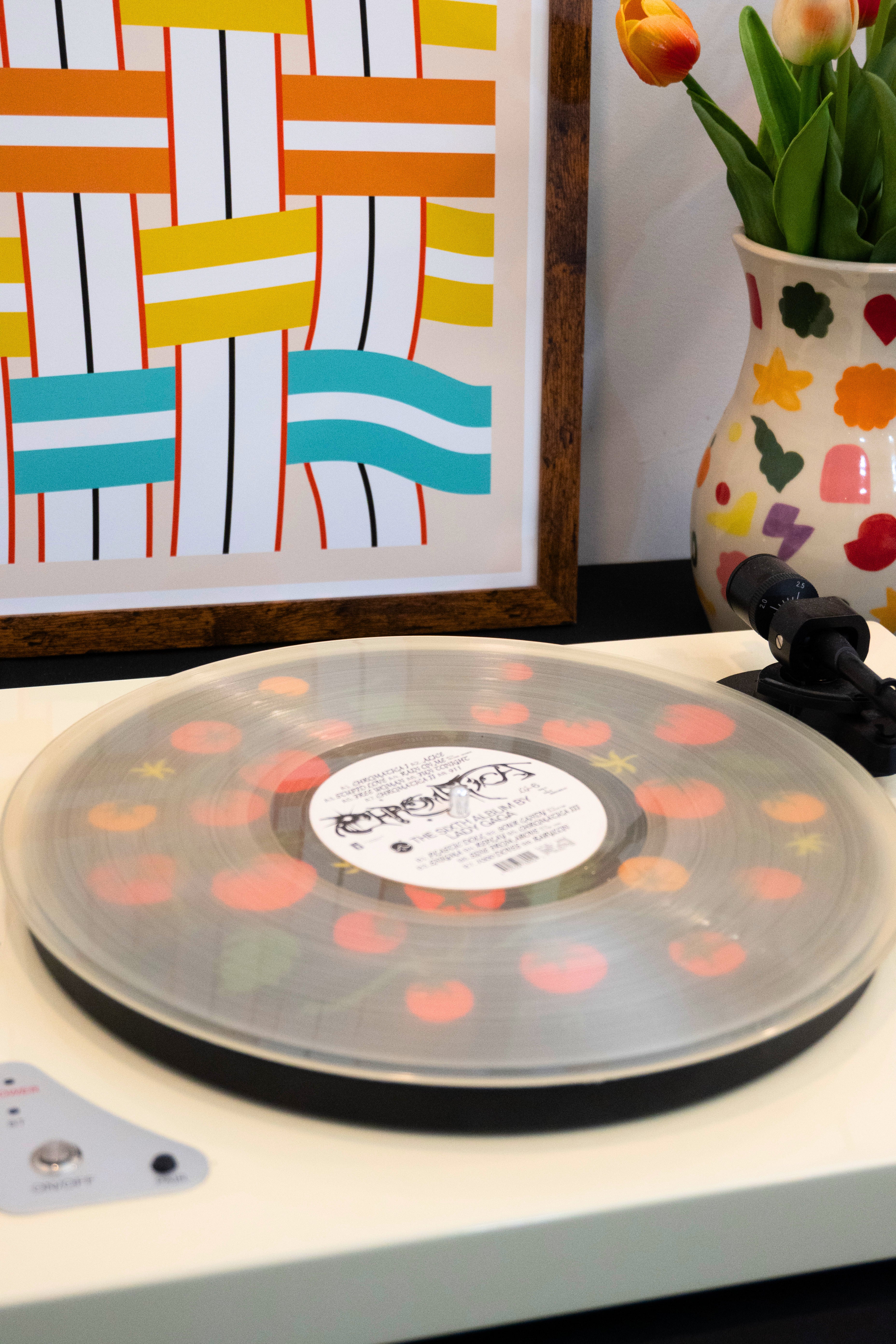 Record player with a transparent vinyl record above a slipmat with a tomato vine design colorful abstract art, and a vase with tulips on a white surface.