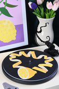 Vinyl record player slipmat with a graphic yellow lemon citrus design on a black background - shown with styled objects and art