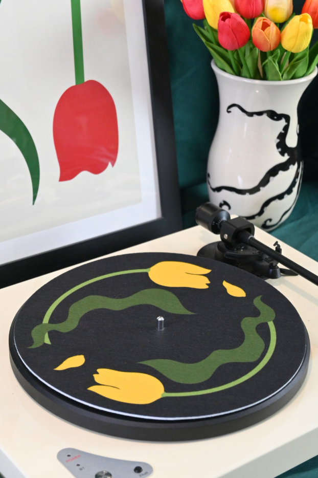 Turntable with yellow tulip with leaves design on black background, framed artwork, and vase of tulips on a table.