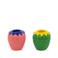 Two strawberry-shaped ceramic candle holders, one pink with yellow spots and blue top, the other green with red spots and yellow top, on a white background.