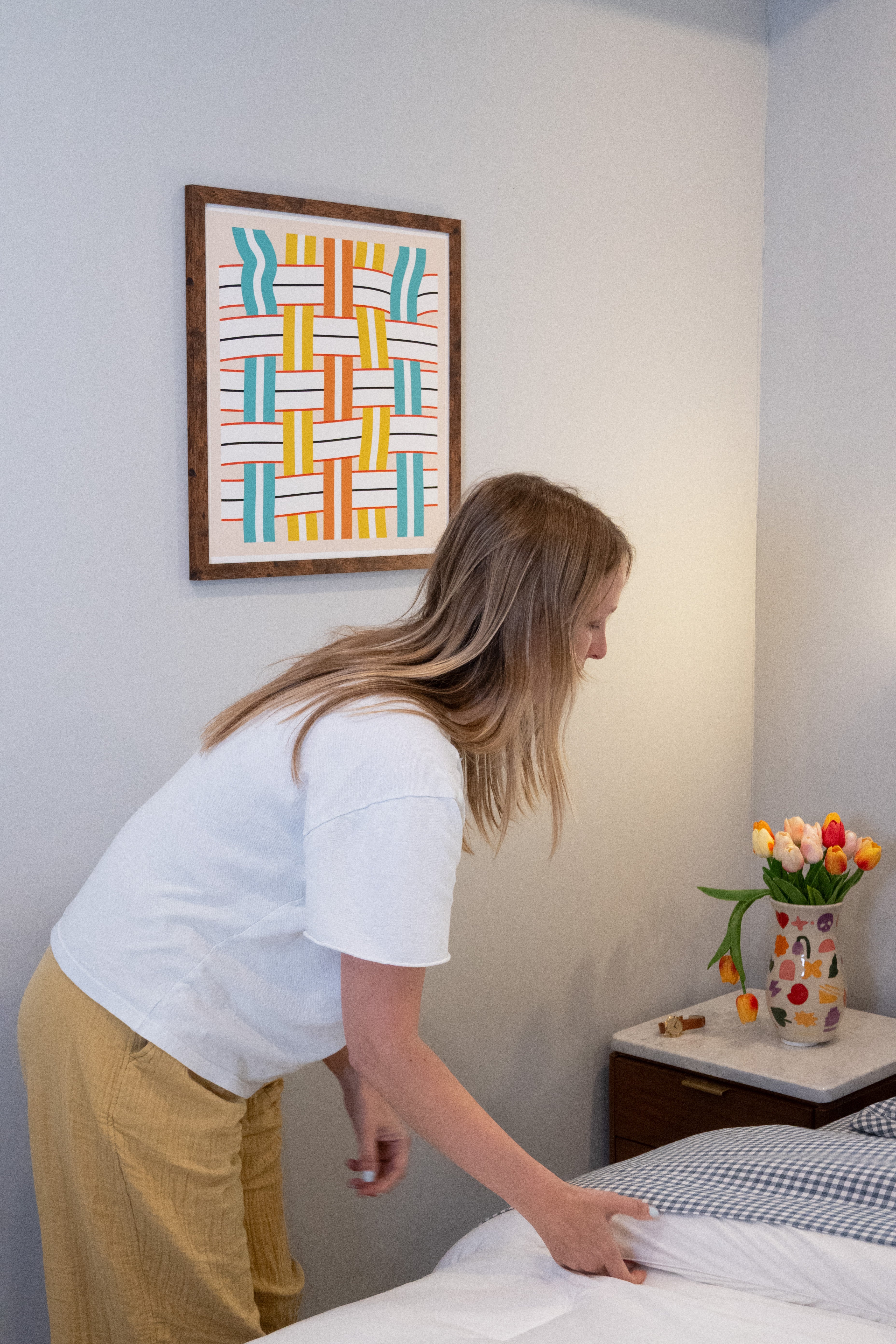 Woman making a bed in a room with a colorful abstract lawn chair pattern painting on the wall.