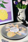 Vinyl record player slipmat with a yellow lemon twist graphic design on a black background - shown beneath a transparent record - styled with artisan objects and art