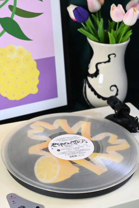 Vinyl record player slipmat with a yellow lemon twist graphic design on a black background - shown beneath a transparent record - styled with artisan objects and art